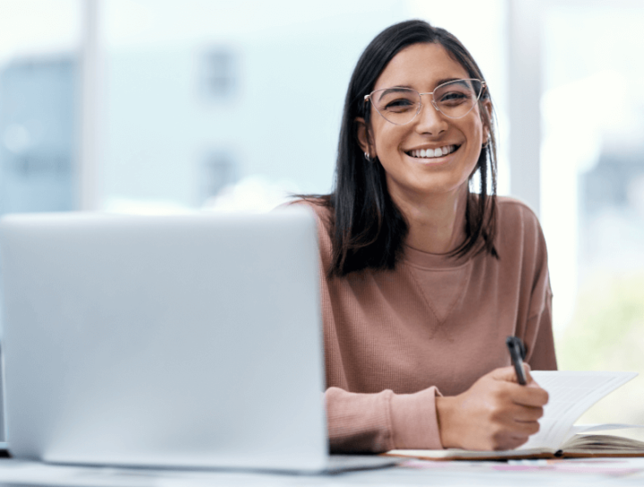Image of a woman holding a pen and paper smiling in front of a laptop