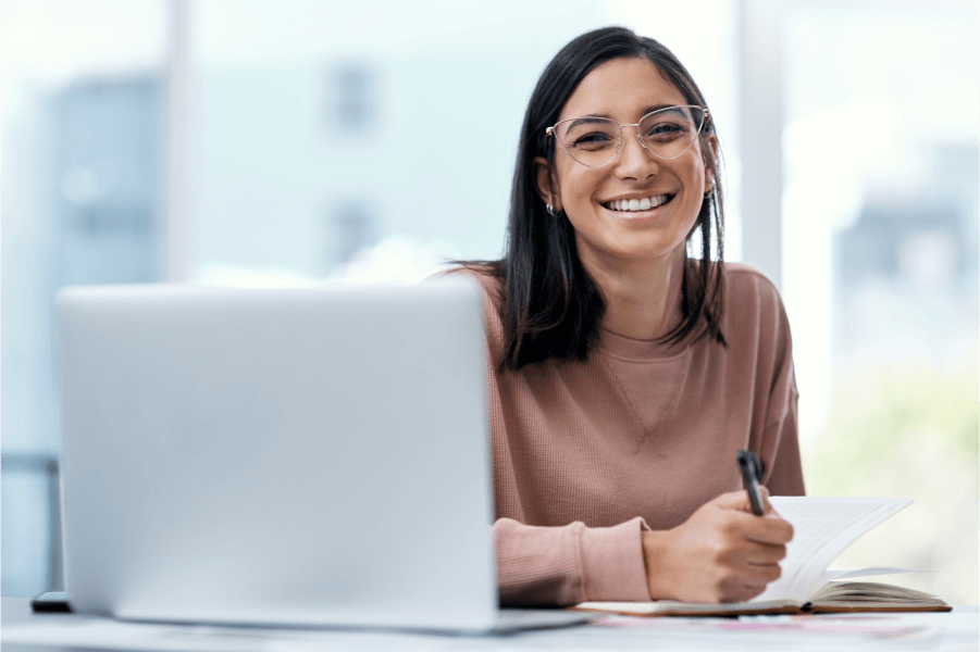 Image of a woman holding a pen and paper smiling in front of a laptop