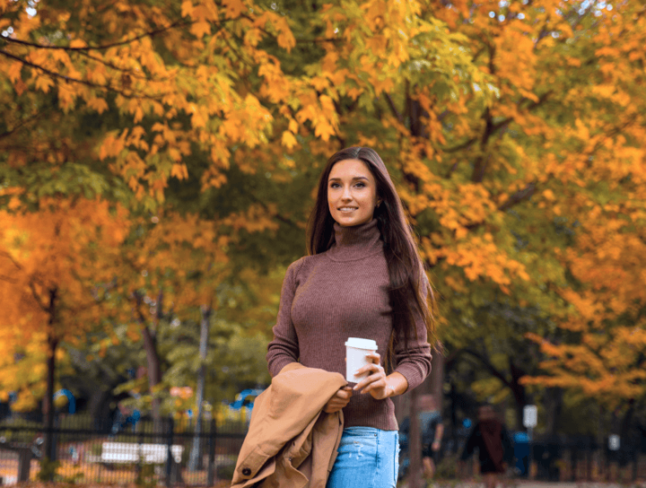 Image of woman with a cup of drink in her hand with autumn trees behind her