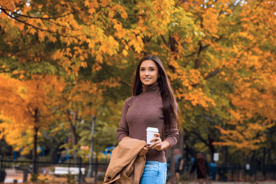 Image of woman with a cup of drink in her hand with autumn trees behind her